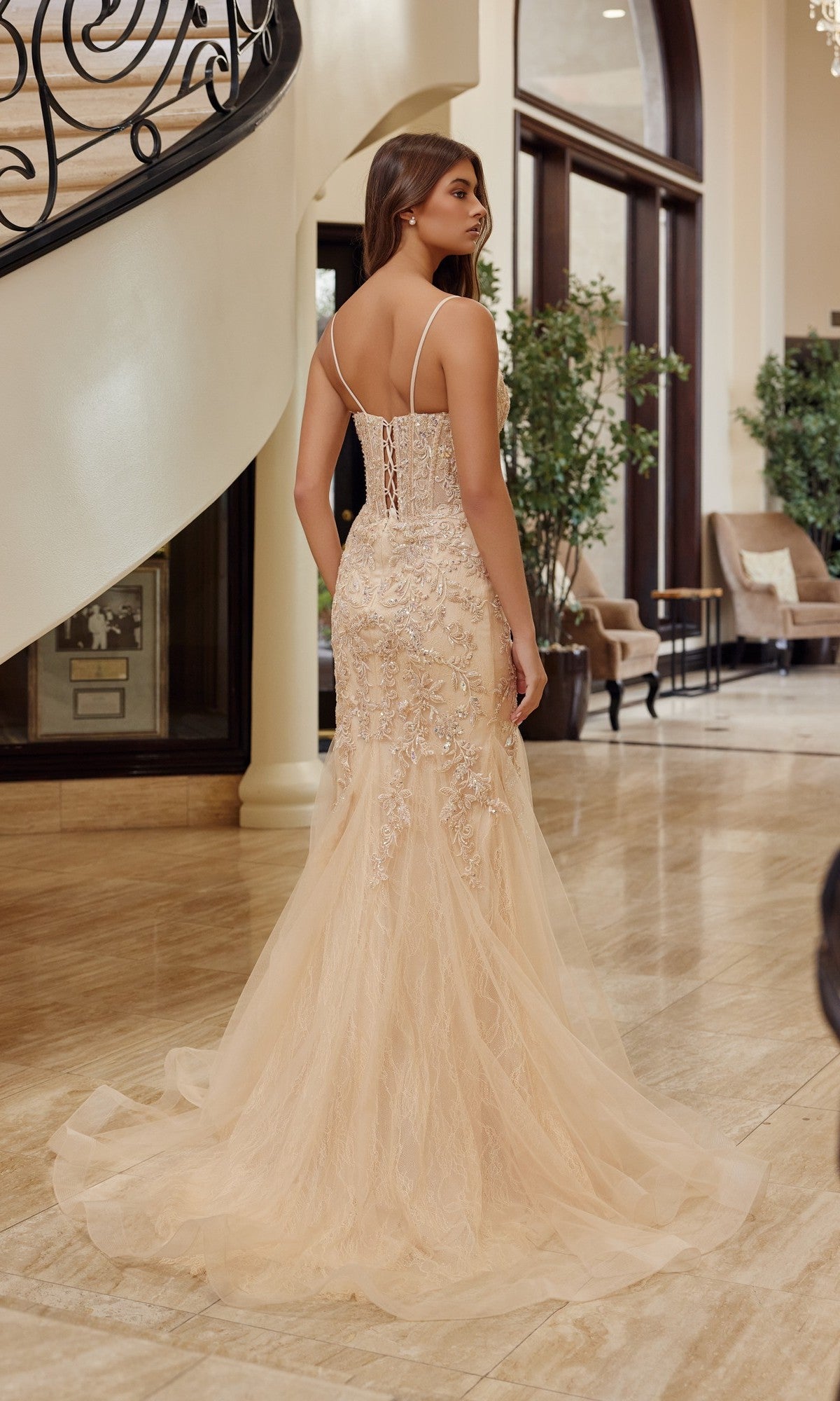 A woman poses indoors near a staircase wearing the Nox Anabel H1874, a fitted floor-length prom dress with lace details, thin straps, a lace-up back, and a subtle trumpet silhouette.